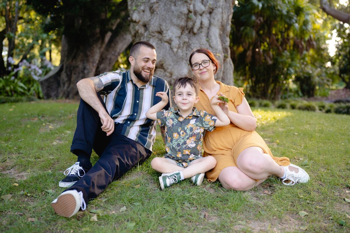 Relaxed family portrait in Geelong with parents and their young son sitting on the grass in front of a large tree during a garden photo session.
