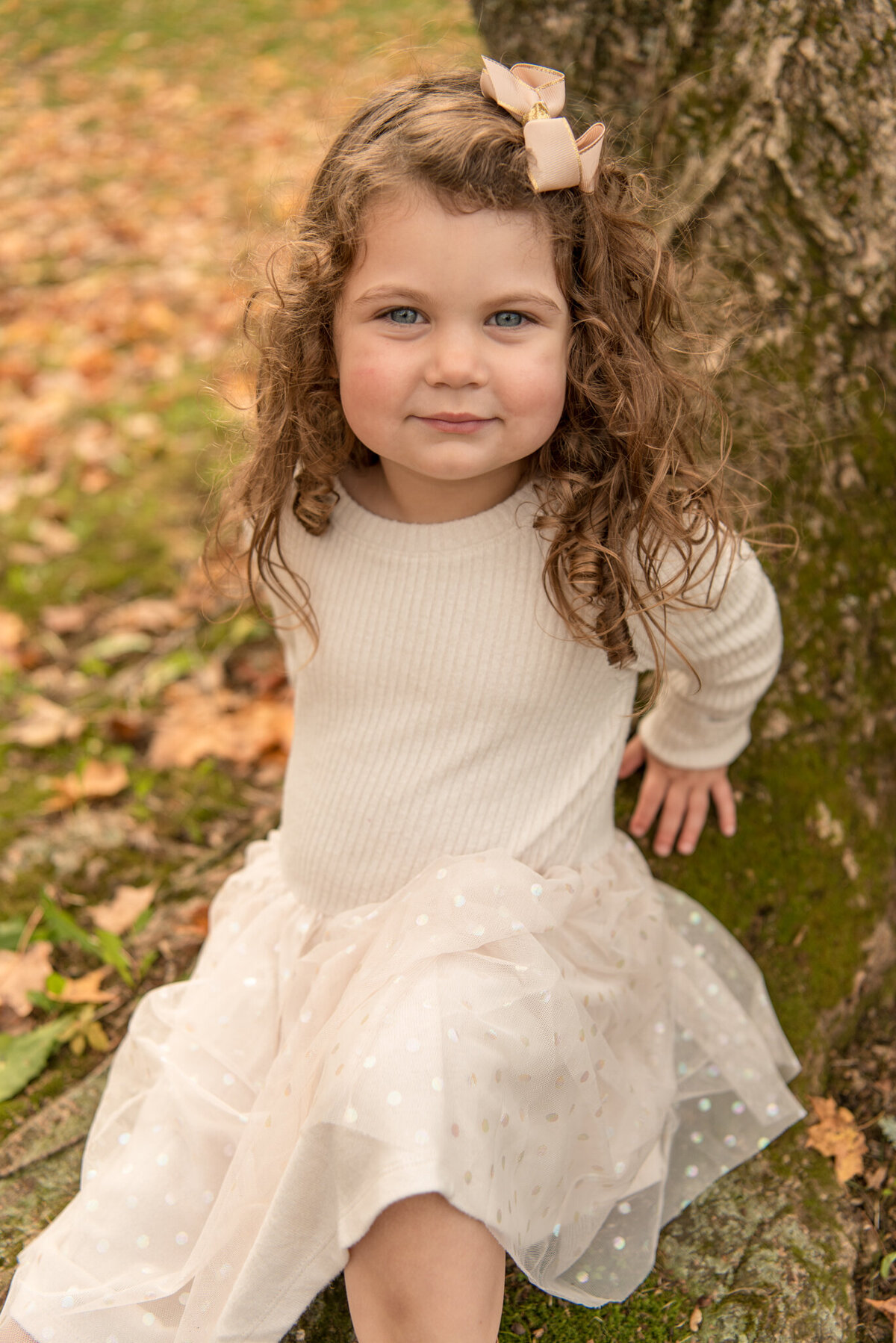 Young girl in white dress sitting at the base of a tree with fall leaves all around
