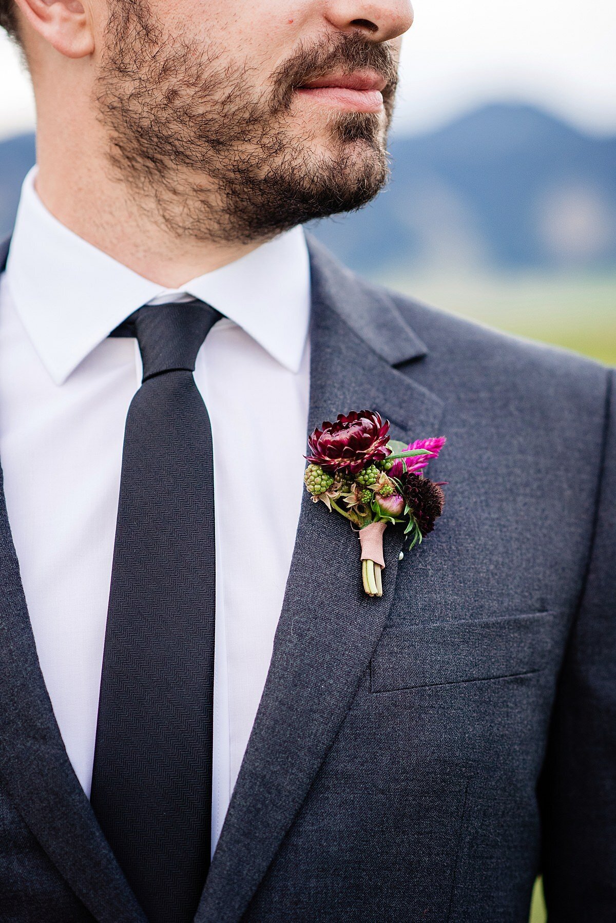 Detail of the groom's boutonniere. The boutonniere is burgundy and pink flowers wrapped in a pink ribbon. The groom is wearing a charcoal jacket with a white shirt and a black tie.