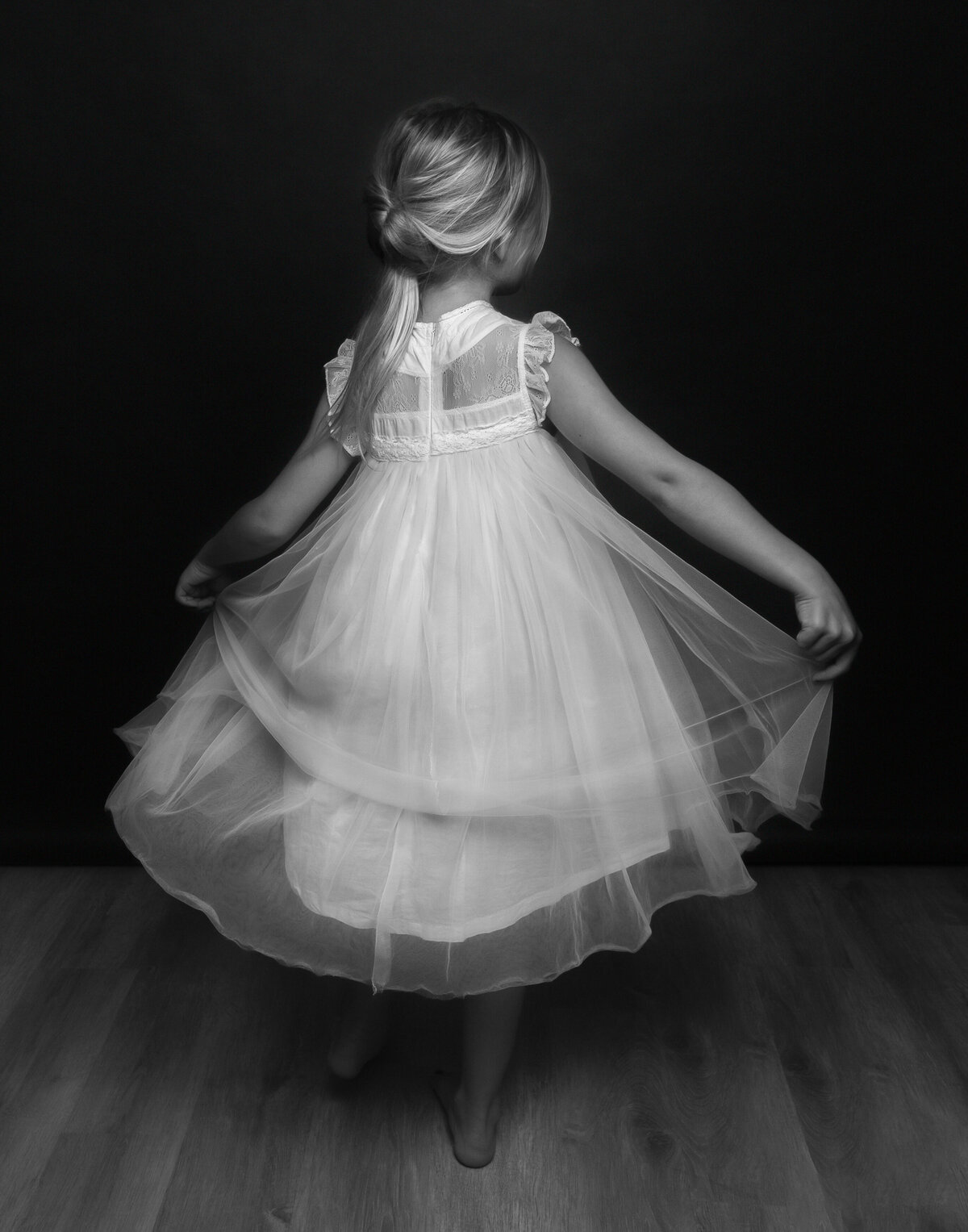 child in flowing satin dress facing backwards with bare feet on studio floor and hands spreading satin dress apart