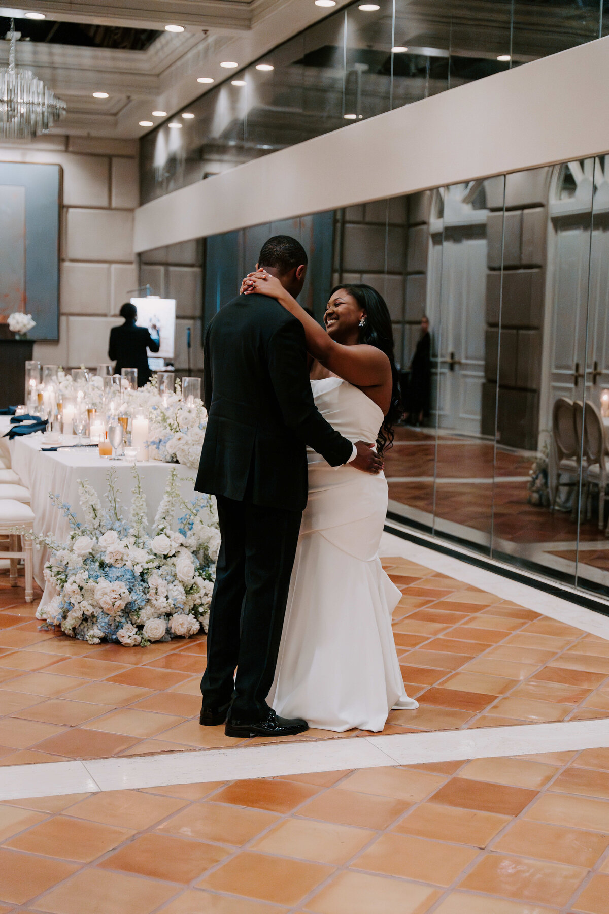 A couple dances elegantly in a warmly lit ballroom. The bride in a white gown smiles joyfully, surrounded by floral arrangements and reflected in mirrored walls.