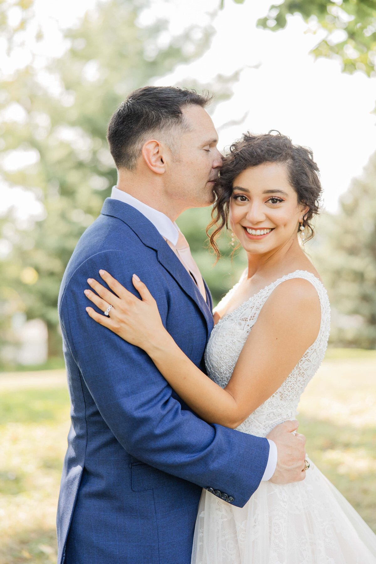 A bride and groom smiling and embracing outdoors in Iowa, the groom kissing the bride's forehead in a sunny park setting.