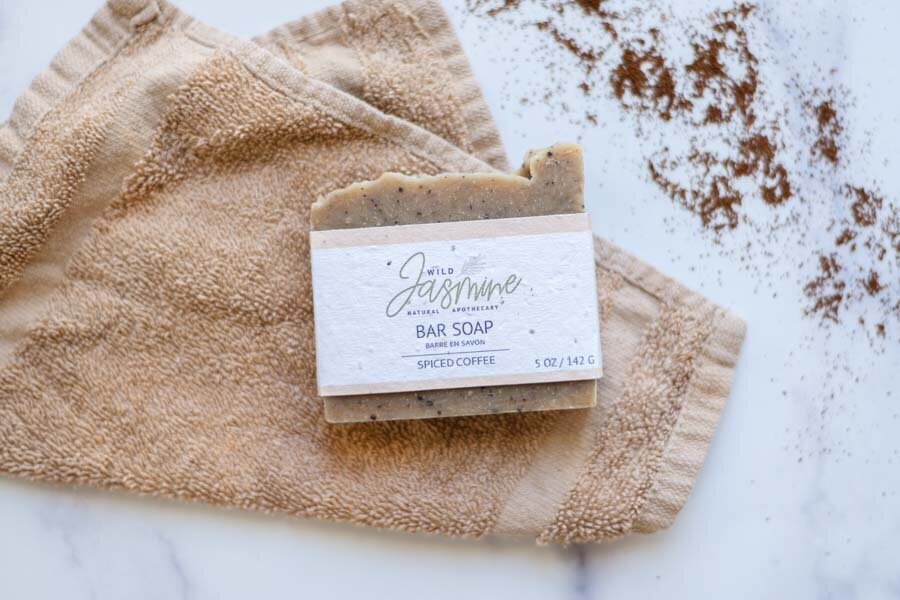 A bar of "jolie jasmine" spiced coffee soap rests on a beige towel, with loose coffee grounds scattered beside it on a marble surface.