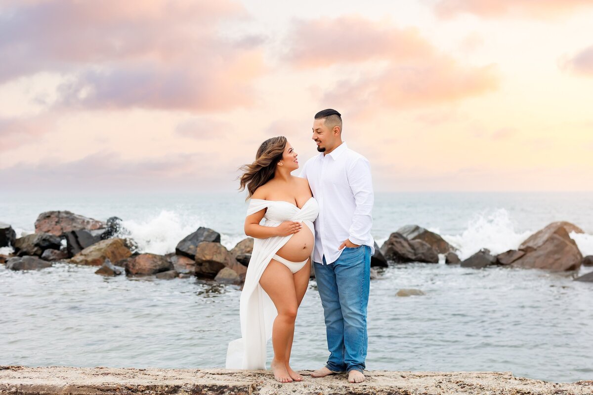 Couple looking at eachother while posing for a maternity portrait in front of the rock jetty at the Hotel Del Coronado with waves splashing around them