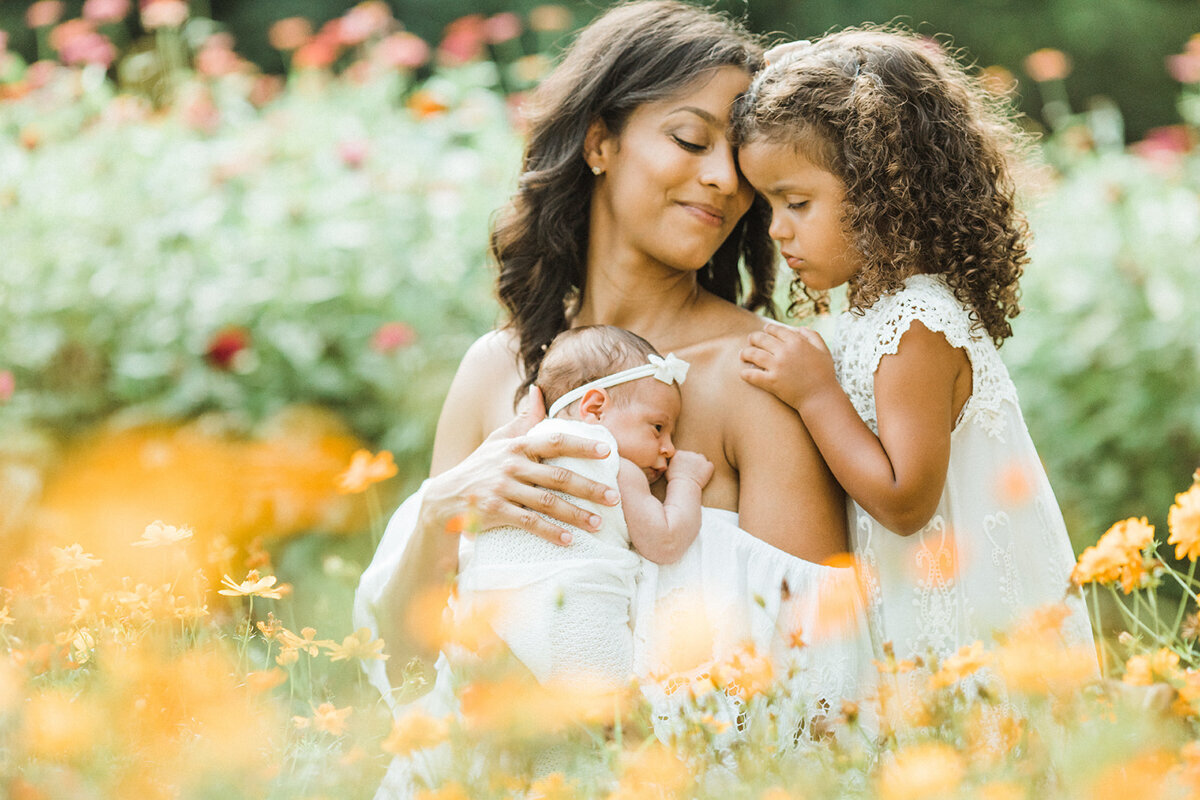 black mom holds newborn close while older daughter hugs mom while standing in a field of flowers