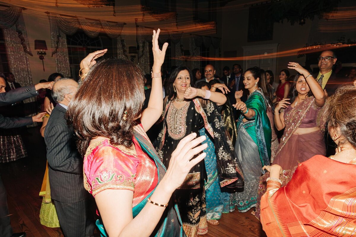 Women in traditional south asian attire dancing joyfully at an indoor celebration.