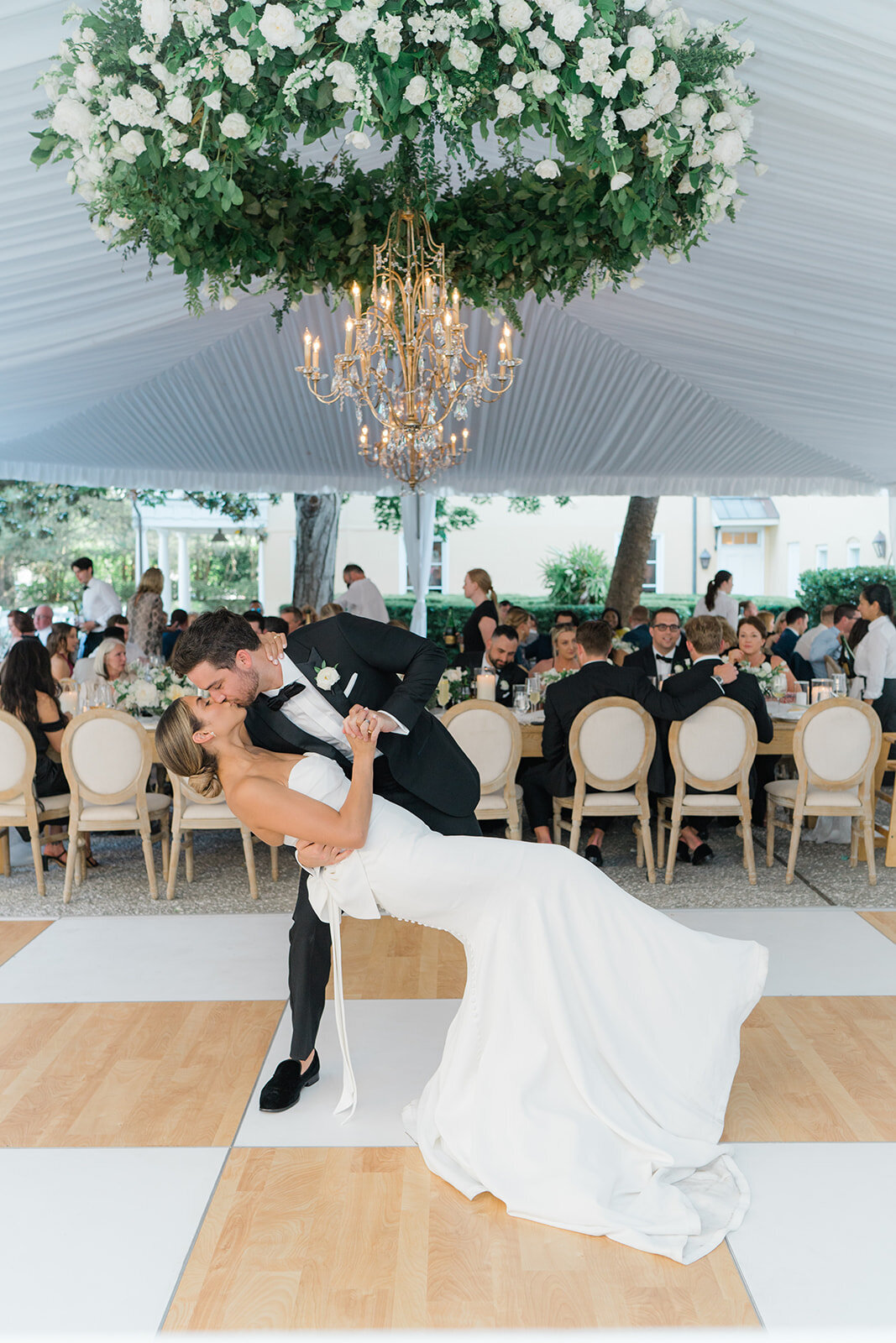 Bride and groom dip kiss on the checkerboard dance floor in draped tent with chandelier circled in white and green flowers. Spring destination wedding in Charleston.