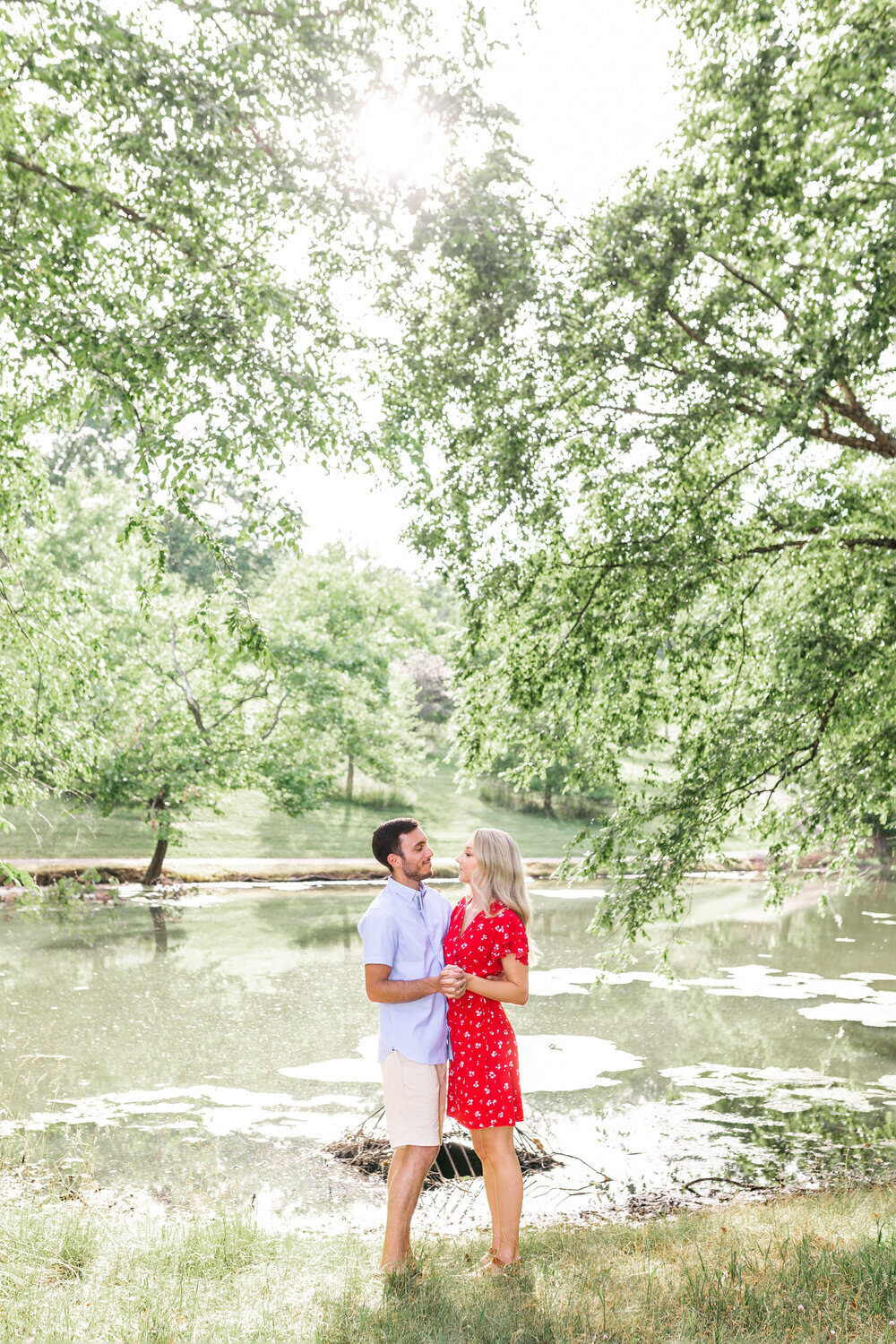 Lady in red dress stands holding hands with fiance by a lake