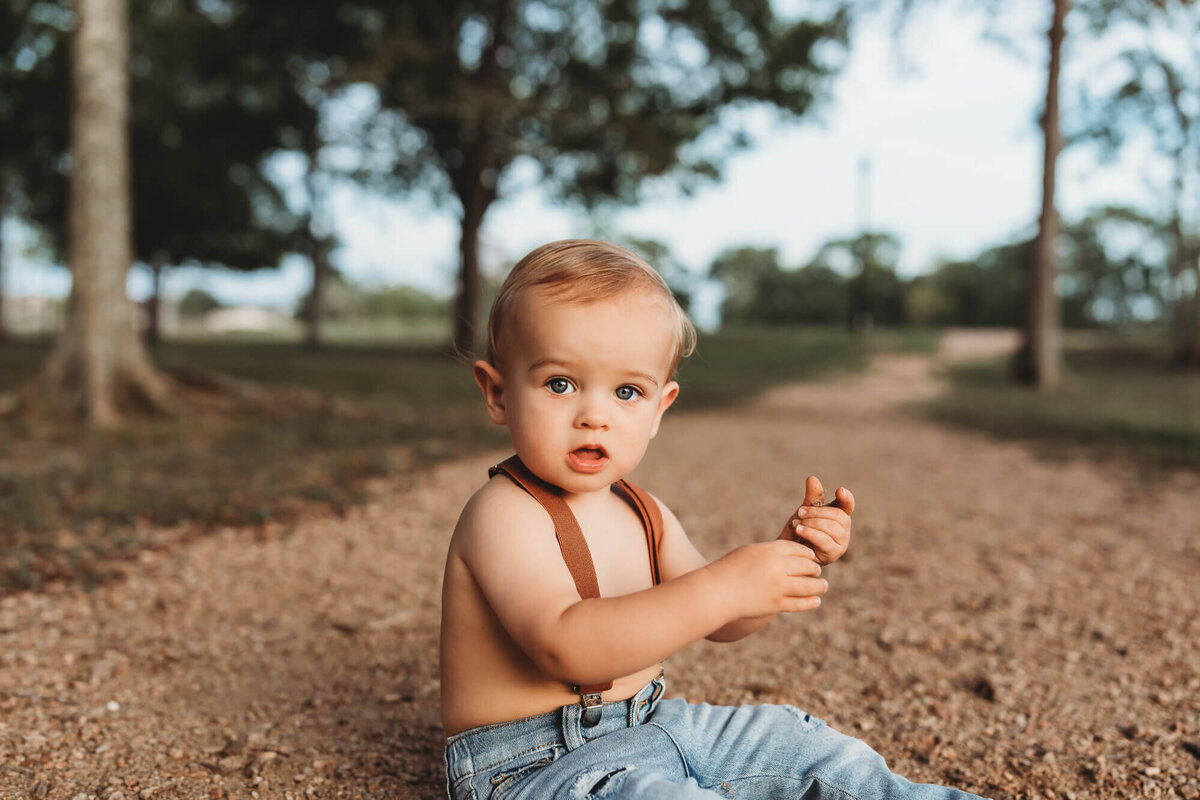 little boy in suspenders sitting on gravel playing with the rocks.
