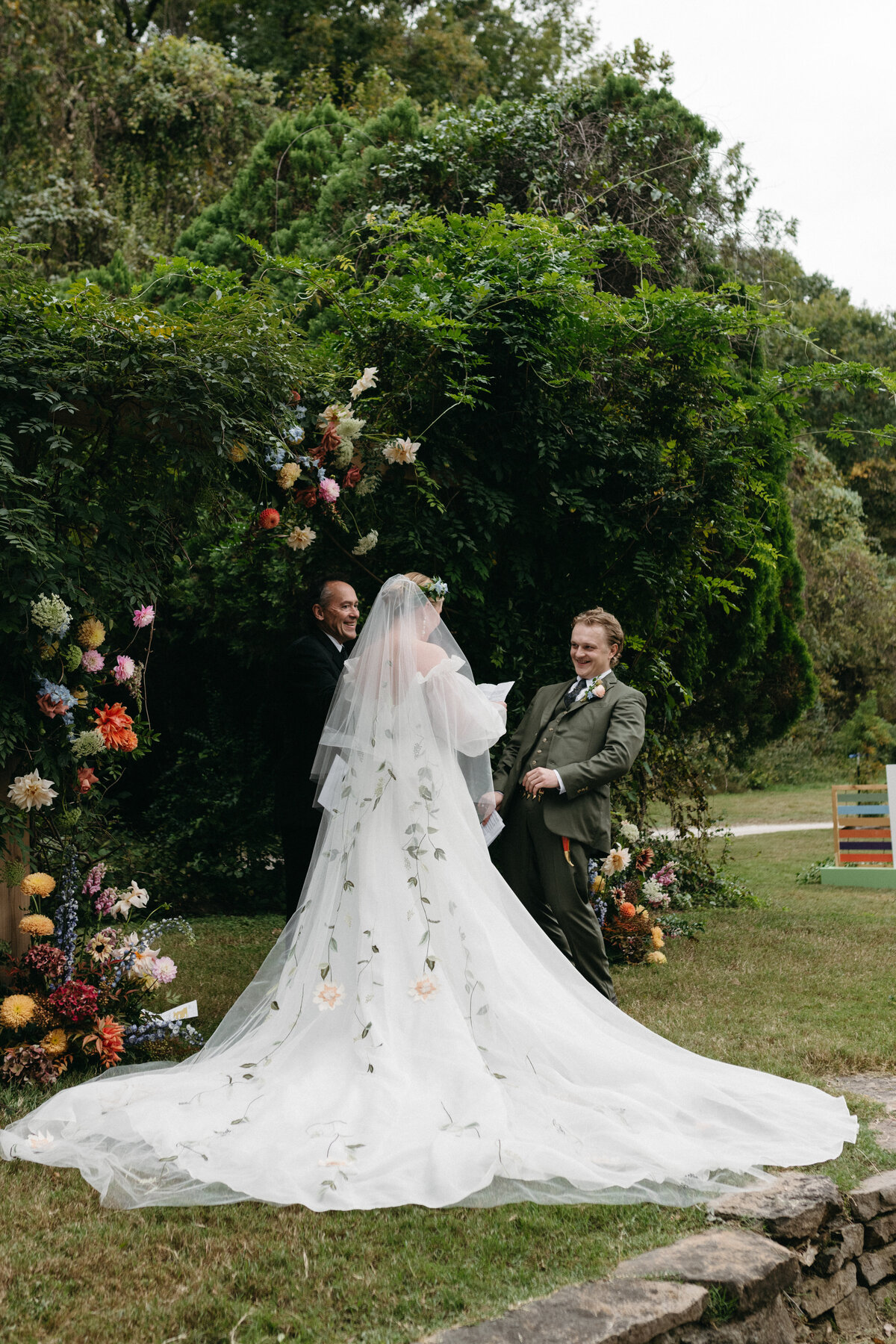 Bride and groom laughing joyfully beneath a lush overgrown ceremony arch embellished with colorful dahlias and garden blooms, capturing a whimsical, romantic moment during their outdoor wedding designed with nature-inspired florals.