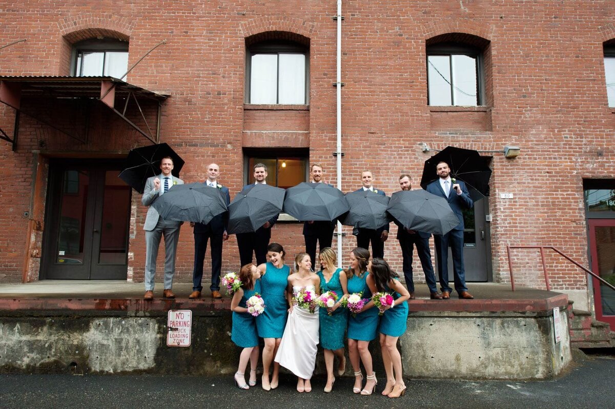 groomsmen standing on a loading dock hold umbrellas over bridesmaids standing on the ground below