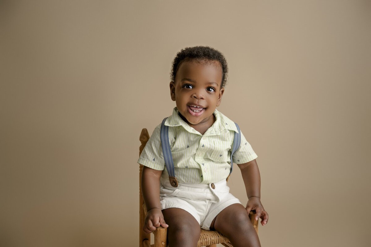 1-year-old boy sits in a chair dressed in a green shirt white shorts and suspenders on a brown background celebrating his first birthday