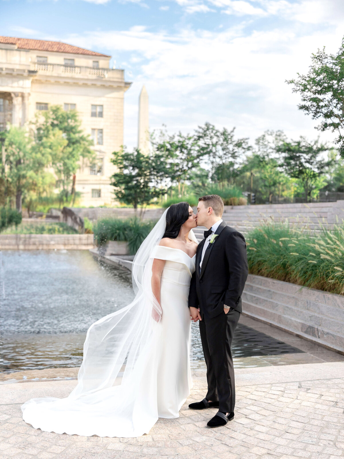 A bride in a white gown and veil kisses a groom in a black tuxedo, standing hand-in-hand by a fountain. Theyre outdoors with a historic building and greenery in the background, under a partly cloudy sky.