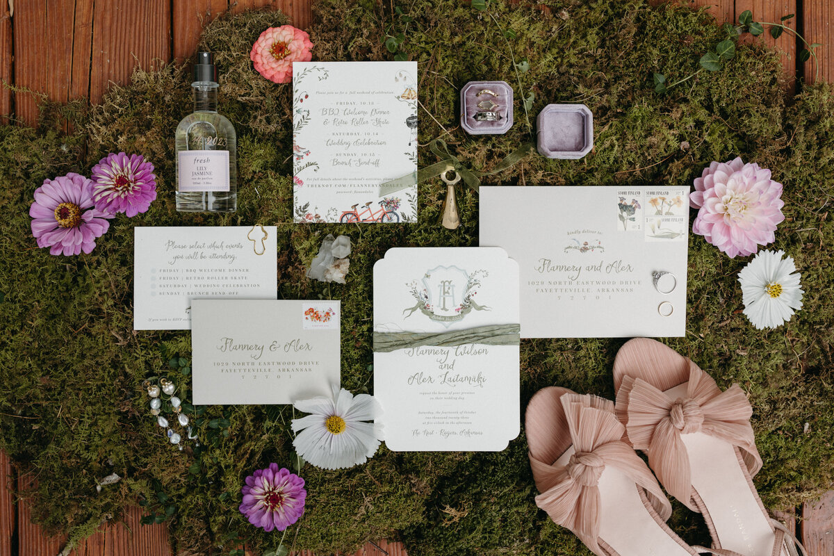 Flatlay of wedding invitation suite styled on natural moss with floral details, custom stationery, blush heels, perfume bottle, vow cards, rings, and fresh seasonal blooms designed by an Arkansas wedding florist.