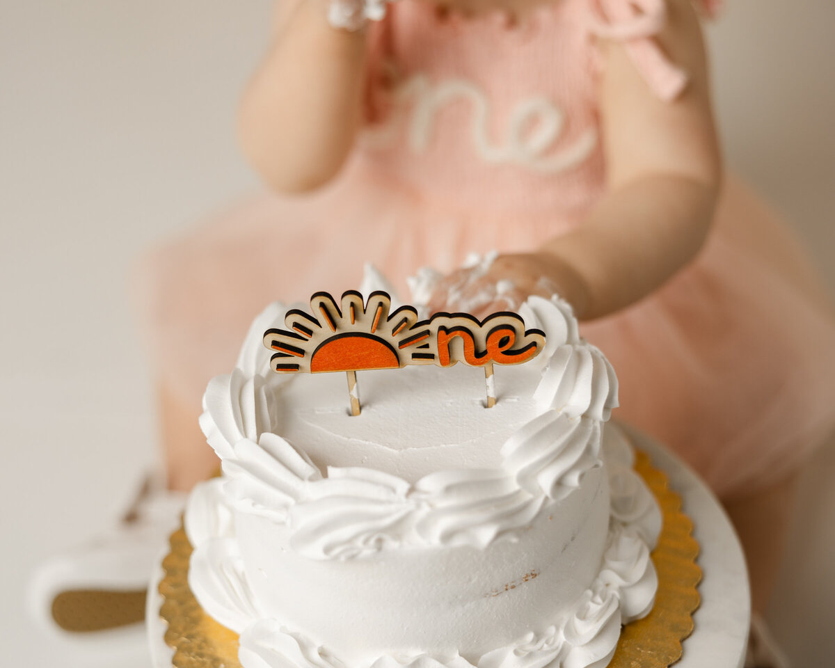 Close-up of a white smash cake with a sunshine “one” topper, as a baby in a pink romper reaches in with frosting-covered fingers.
Photograph by Yucaipa cake smash photographer Kaitlyn Dawn Photography.