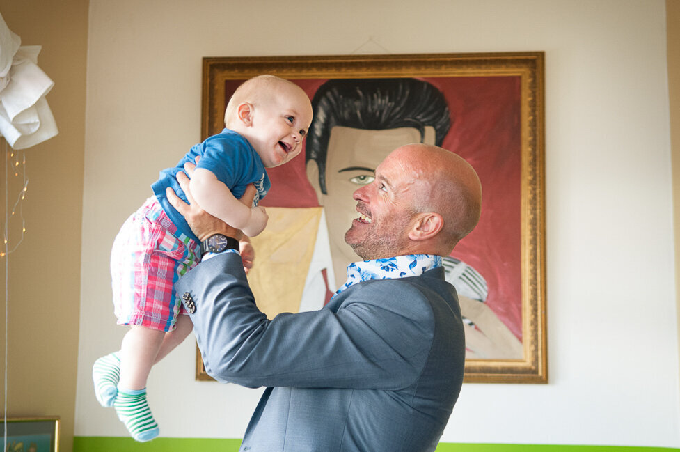 groom wearing a light blue jacket and floral shirt holding his baby son in the air laughing in front of an Elvis portrait in Inch beach cafe