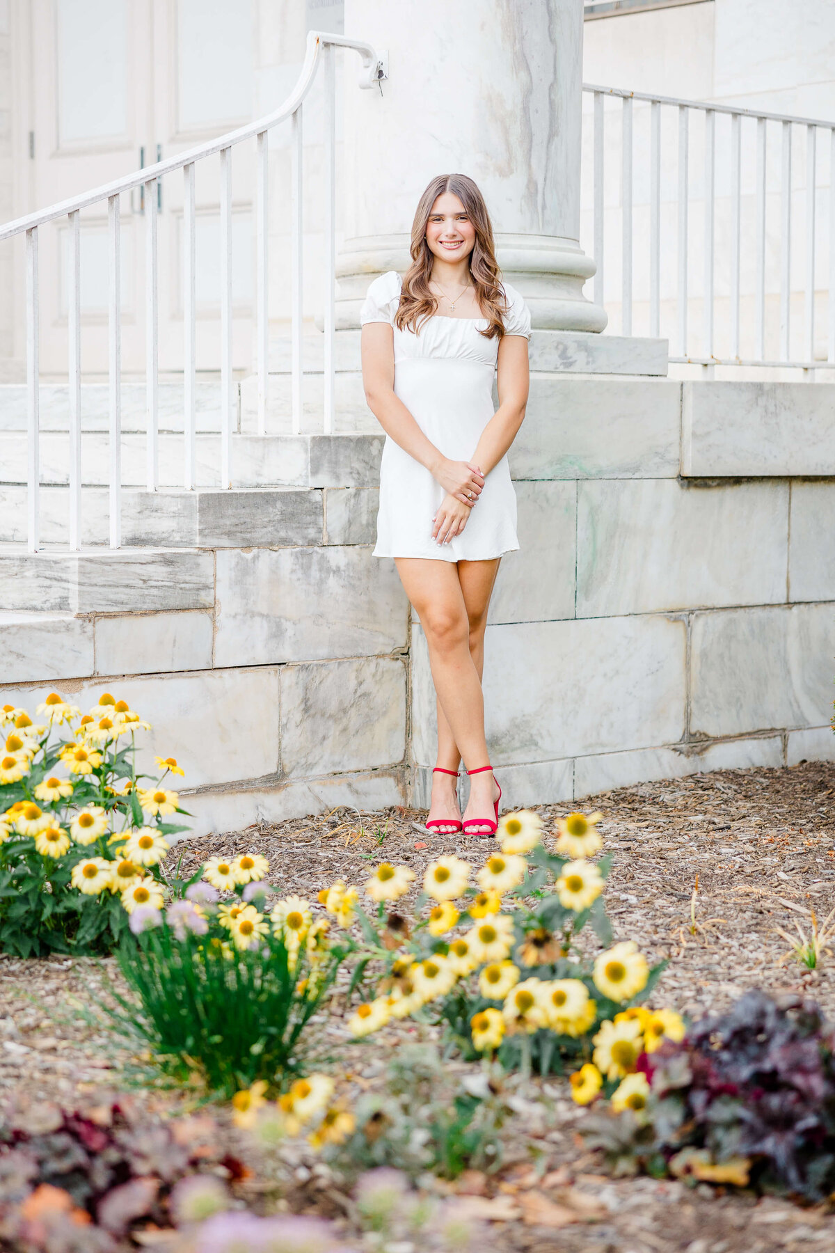 A person in a white dress and red sandals poses gracefully against a marble structure. Yellow flowers blossom in the foreground, framed by a white railing behind them—a perfect composition for a senior portrait photographer Fayetteville NC treasures.