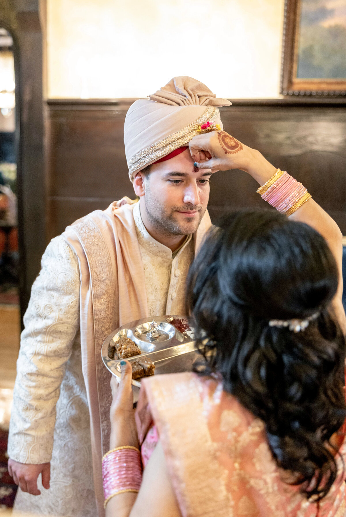 The mother of the bride performs a traditional ritual, placing a red mark on the grooms forehead with ceremonial items on a silver tray. The groom wears a beige sherwani and turban. The mother wears a pink and gold outfit with bangles.
