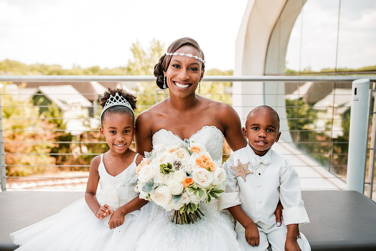 African American bride wearing a strapless wedding dress and rhinestone headband holding a large bouquet of white and orange roses sited in a window at Noah Liff Opera Center with the flower girl wearing a white dress with a rhinestone tiara and the ring bearer wearing a white tuxedo and sheriff badge.