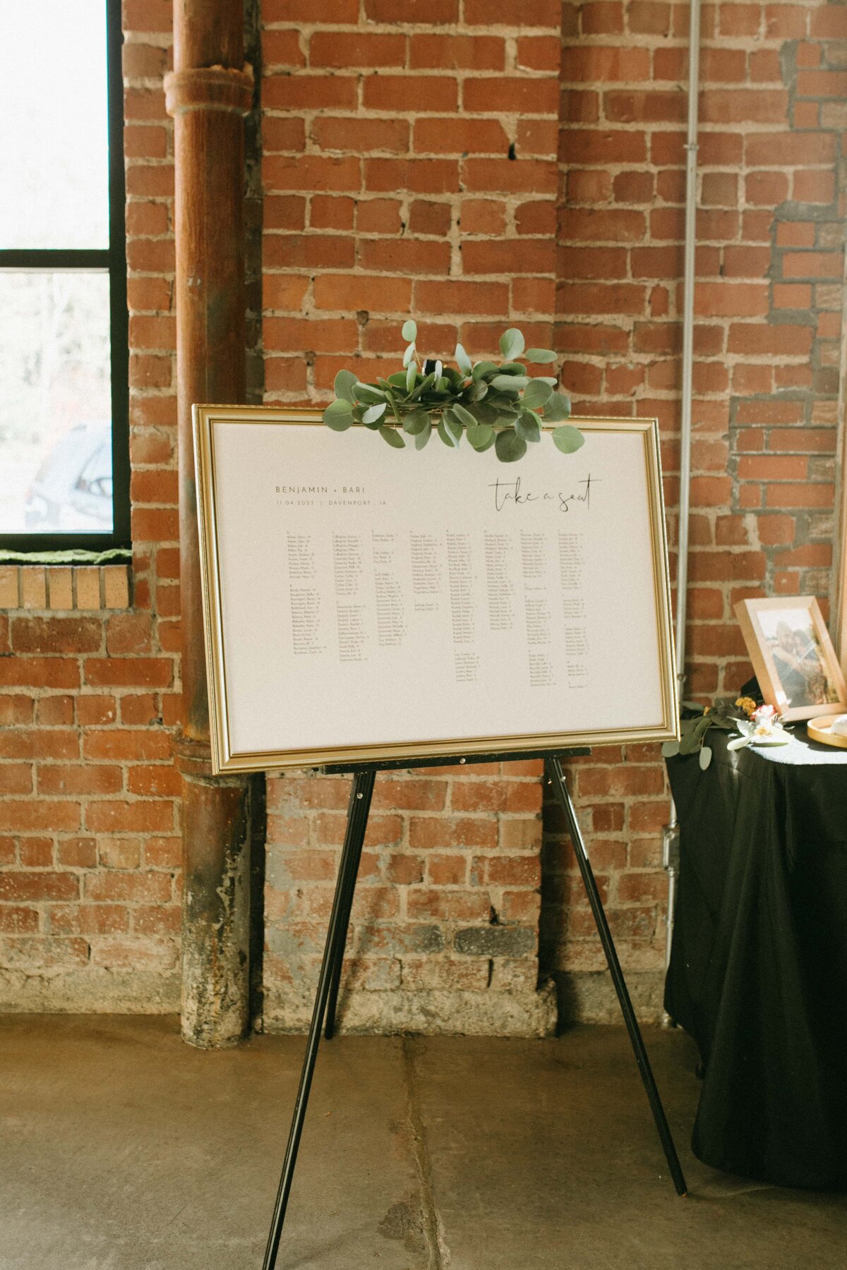 A framed seating chart on an easel at a rustic Iowa event venue, with exposed brick walls in the background.