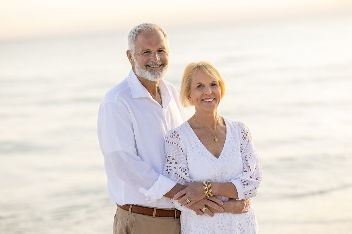 A mature couple smiling with their arms around each other on the beach.