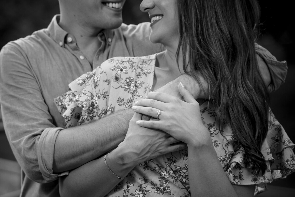Black-and-white-up-close-image-of-the-hands-and-smiles-of-an-engaged-couple-that-is-looking-at-one-other-with-their-male’s-arms-over-the-female’s-shoulders-highlighting-the-engagement-ring