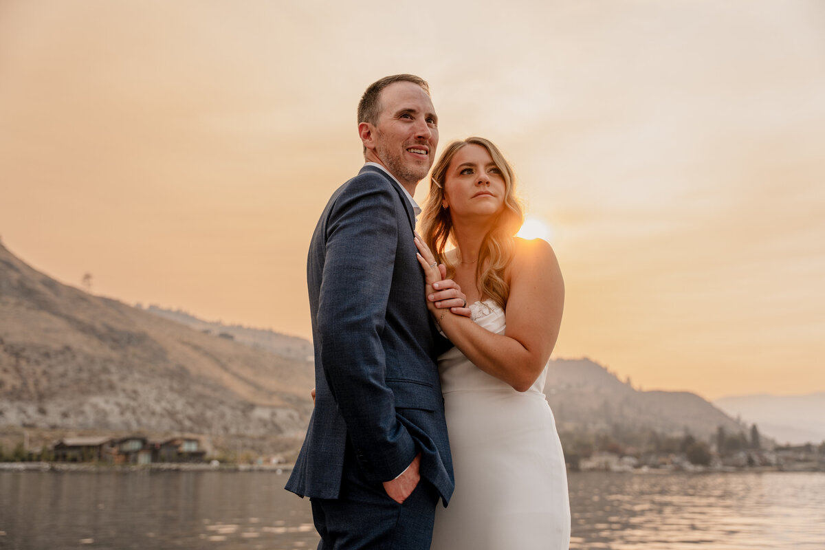 The bride and groom look out at the water during the sunset on a smoky day in Lake Chelan.
