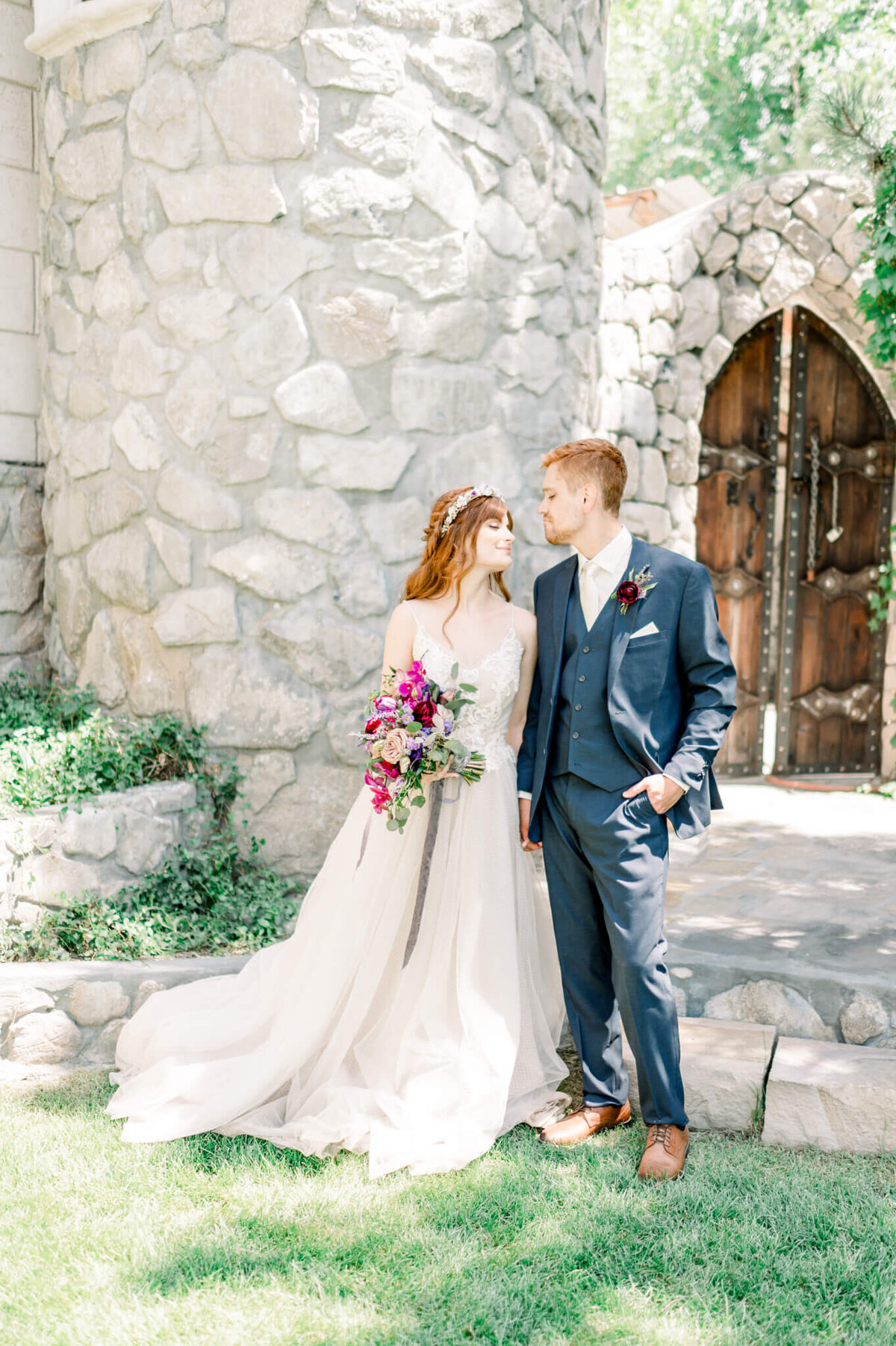 red haired bride and groom in white gown and navy suit hold bright pink flowers and look at one another