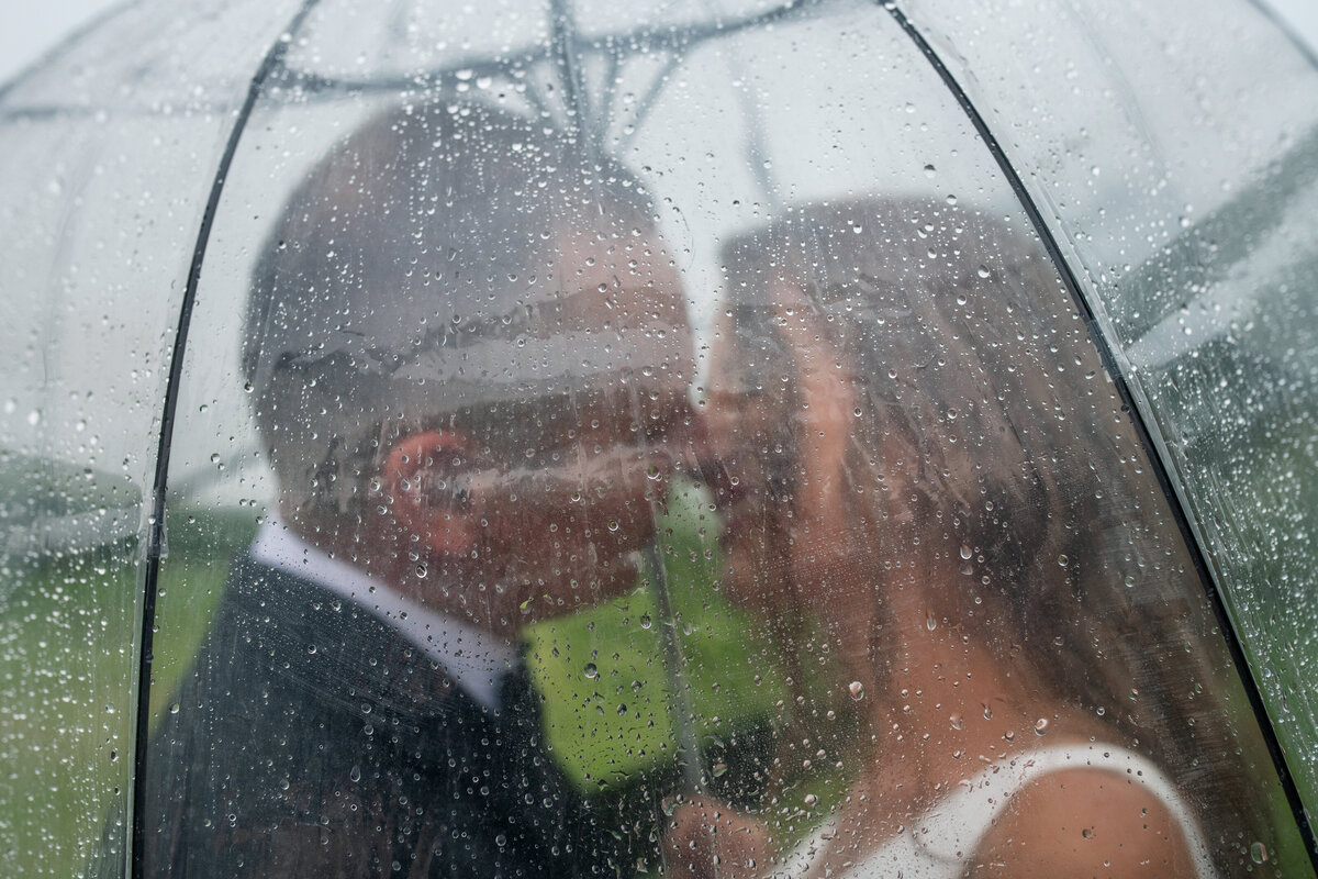 bride and groom on their rainy wedding day kissing under clear umbrella