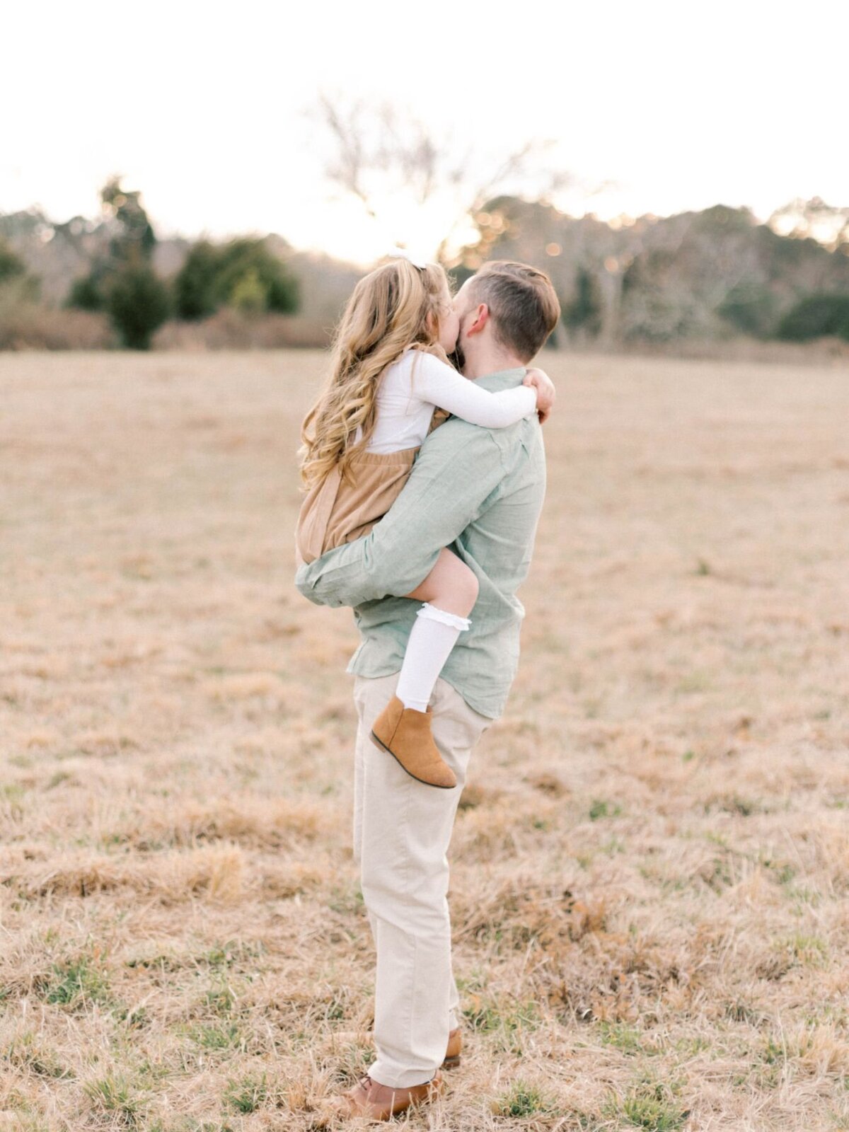 Father holds daughter in is arms in grass field.