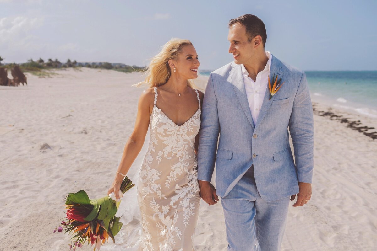 Bride and groom walk down beach holding hands in Cancun