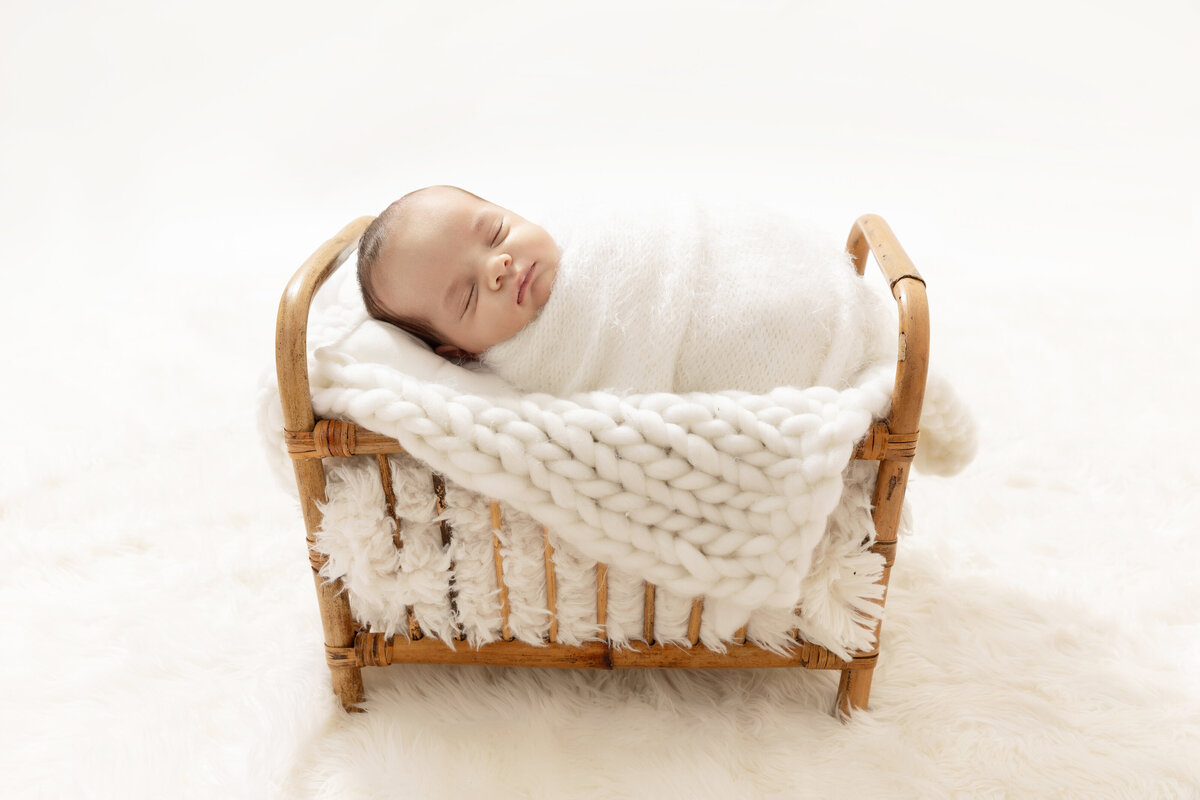 A newborn baby sleeps in a tiny wicker bed in a white swaddle