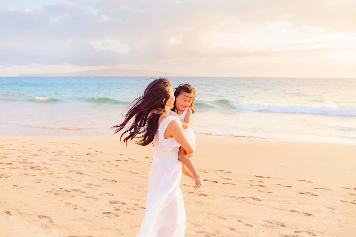 Woman with long back hair holds her toddler daughter on her hip and spins her at sunset as the pair have a fun family photoshoot on Maui with Love + Water