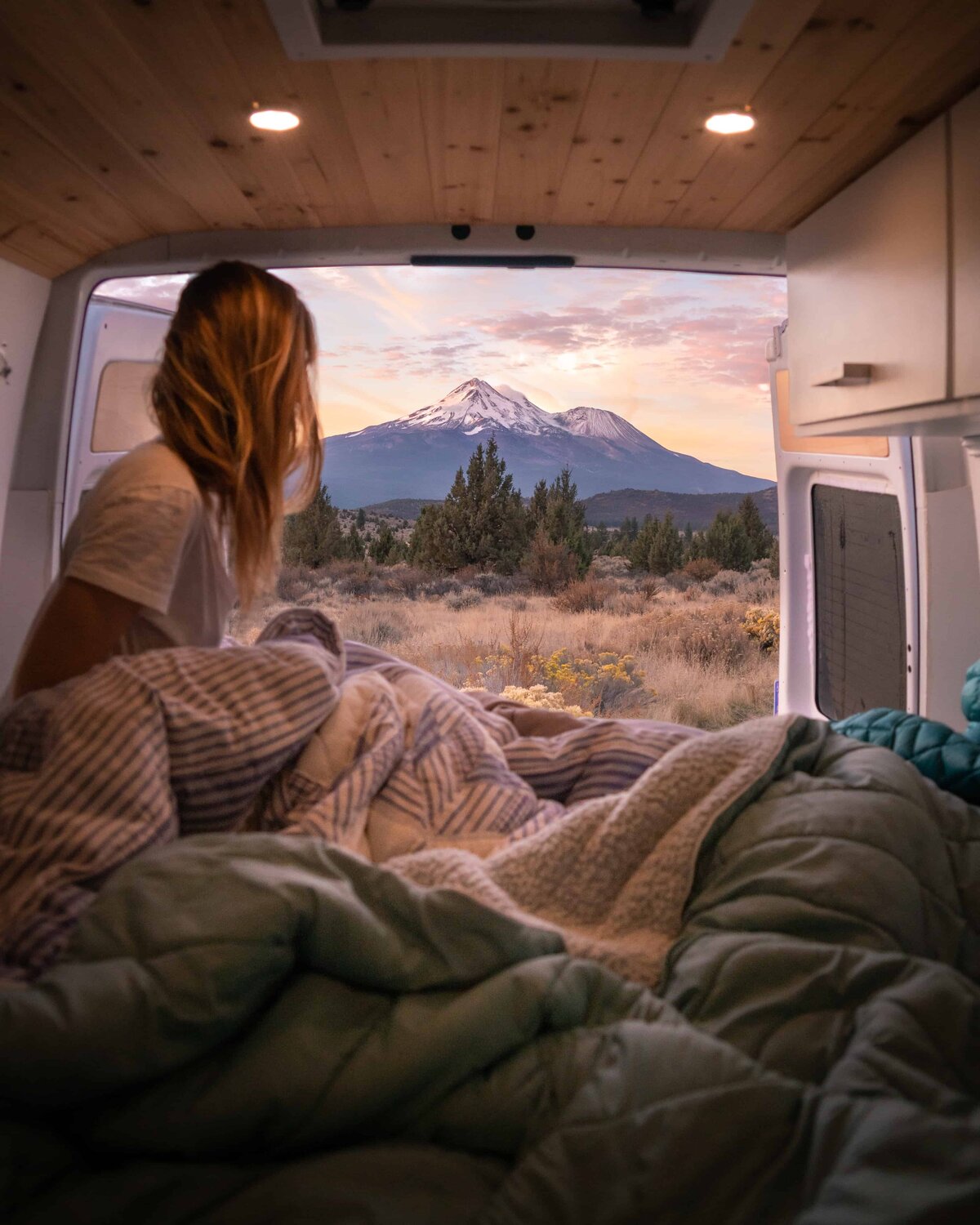 Woman sitting inside a van under blankets looking out at a mountain