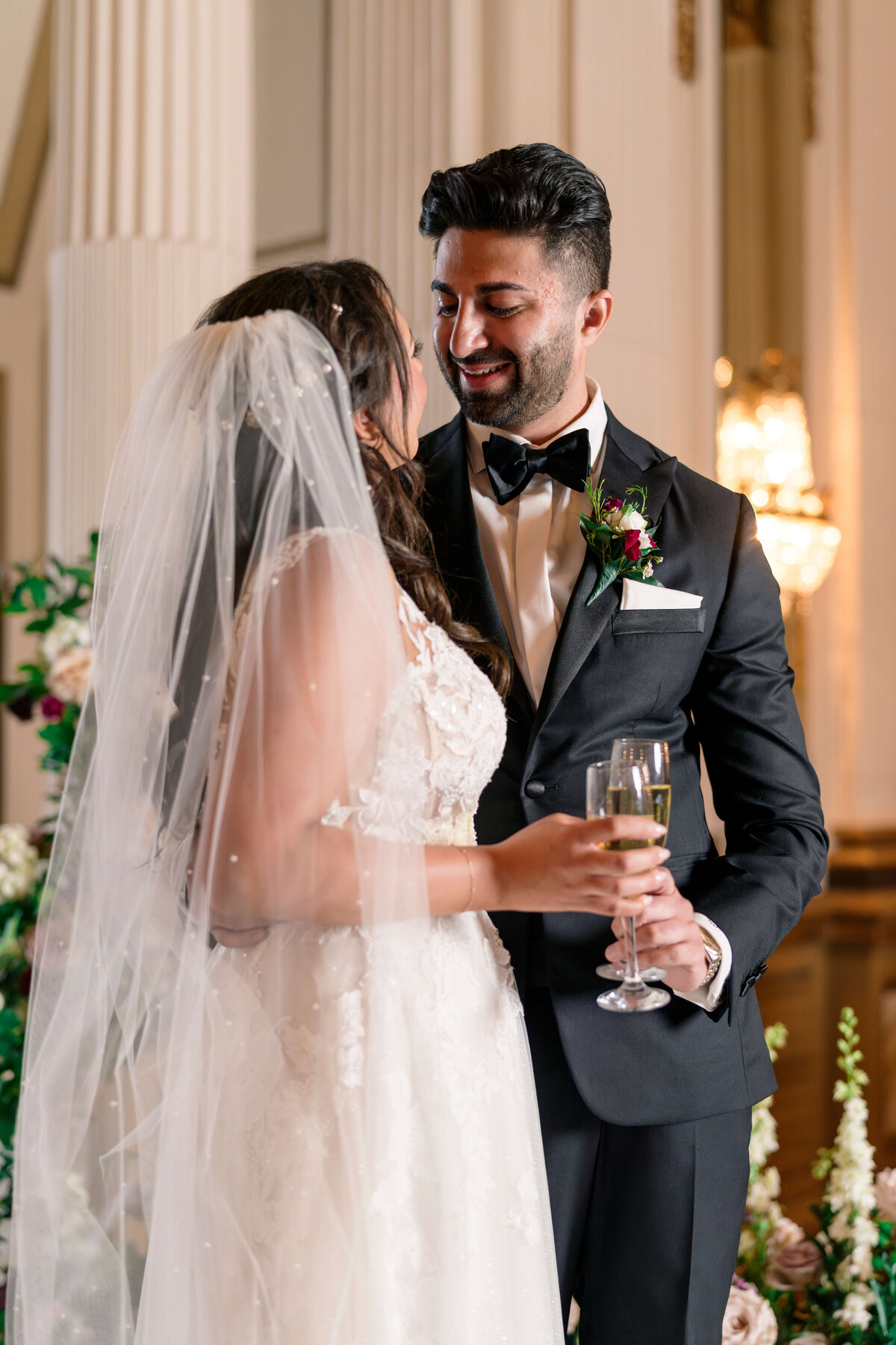 A bride and groom smile at each other during their wedding. The bride wears a white gown and holds a champagne glass, while the groom is in a black tuxedo with a bow tie. They stand in an elegantly decorated venue.