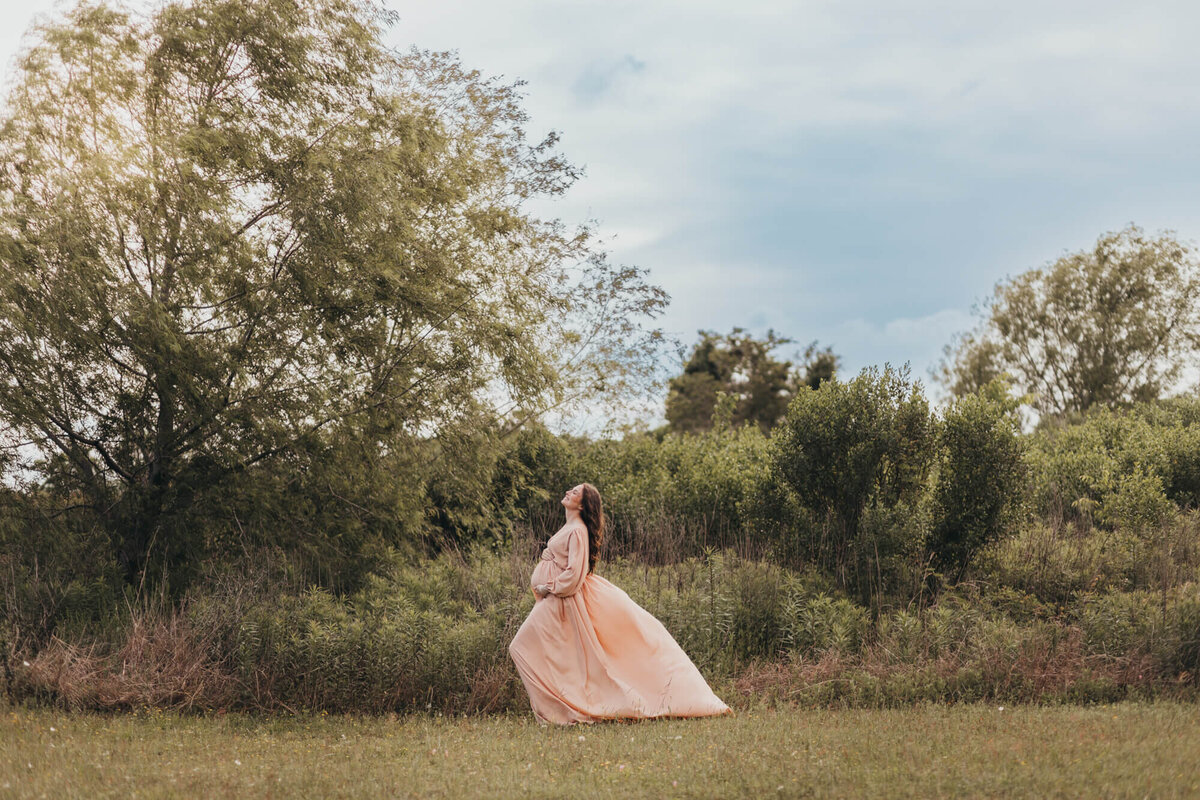 wide open shot of woman standing in breeze for her maternity photos wearing coral colored gown.