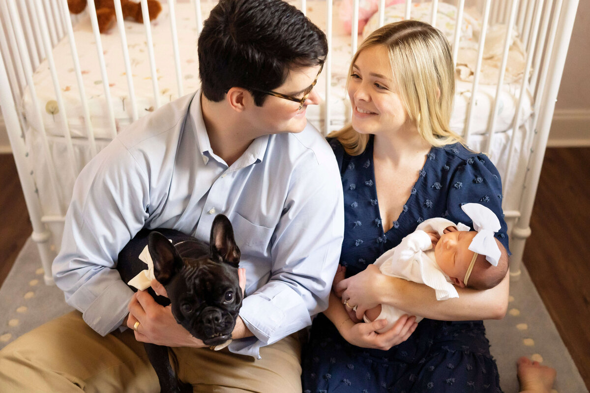 Parents sitting in front of crib with newborn girl and dog
