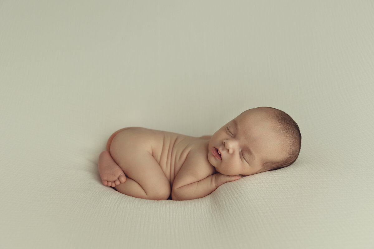 A newborn baby sleeps in froggy pose wearing nothing in a studio