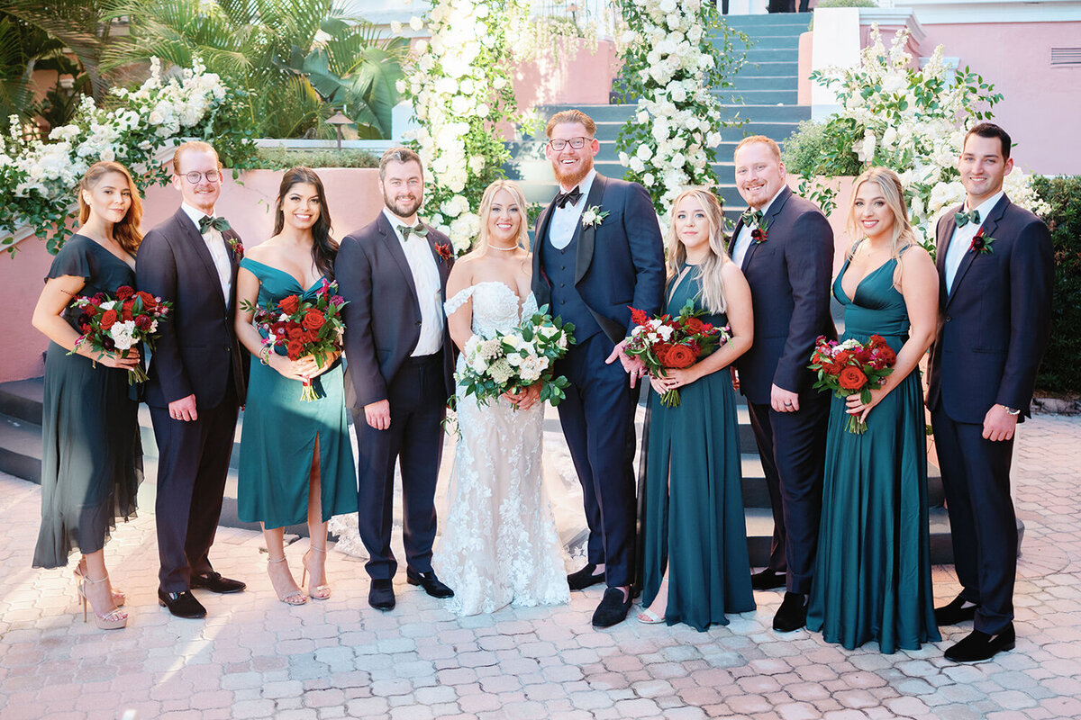 Wedding party in front of a floral arch in the courtyard of Don Cesar Hotel, St. Pete Beach, FL, for a December wedding. Bridesmaids in emerald green satin gowns with red and white bouquets, and groomsmen in navy blue tuxedos with emerald green bowties and red boutonnieres.