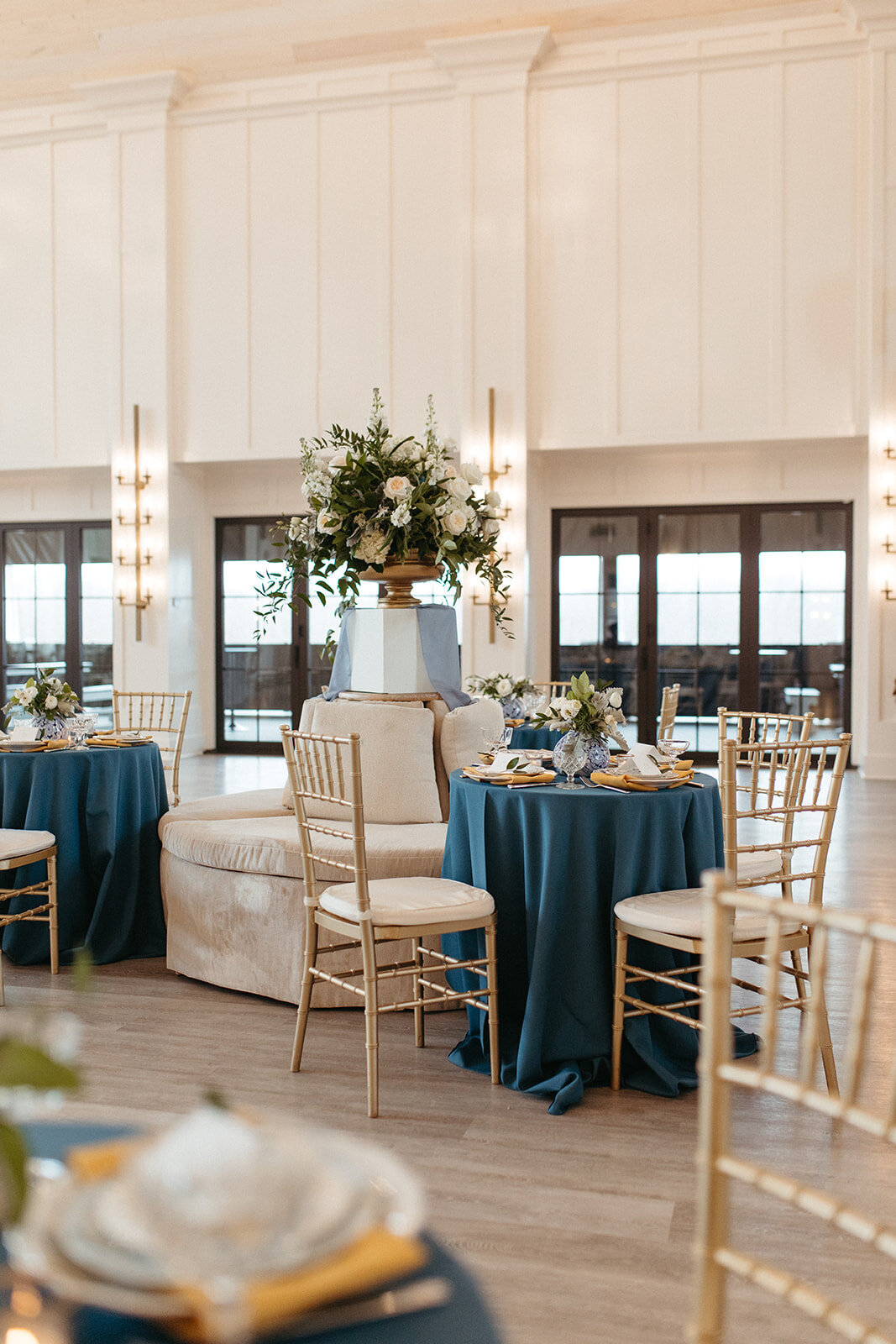 A bright and airy banquet room with blue satin linens on round tables, gold chairs and white floral arrangements.