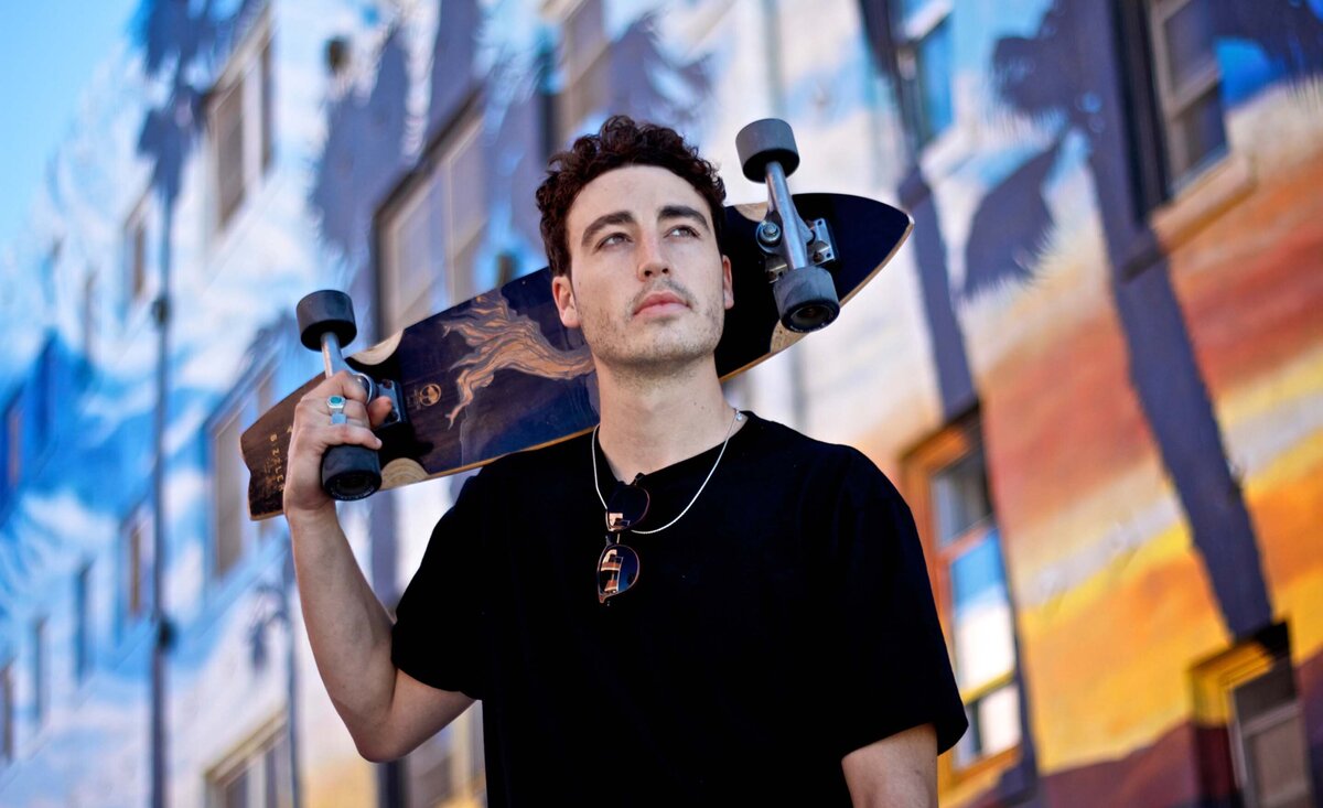 Male musician portrait wearing black shirt with skateboard resting on shoulder palm tree graffiti behind