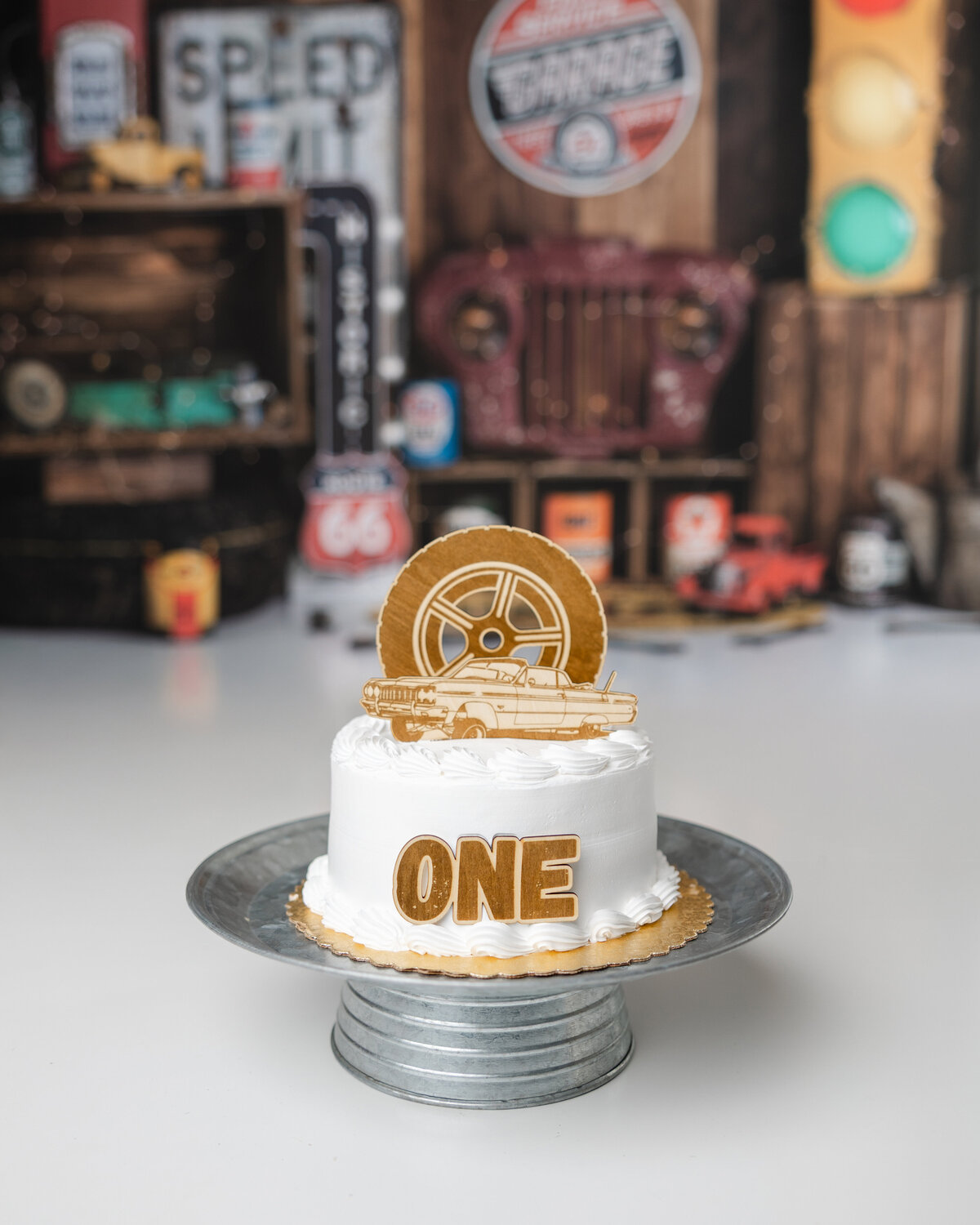 White frosted smash cake with a gold “ONE” topper and vintage car design, set against a garage-themed birthday backdrop.
Photograph by Yucaipa cake smash photographer Kaitlyn Dawn Photography.