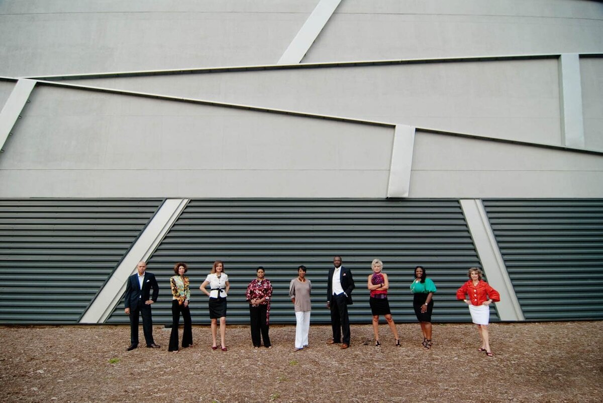 9 coworkers stand together in a horizontal line up in front of modern architecture.