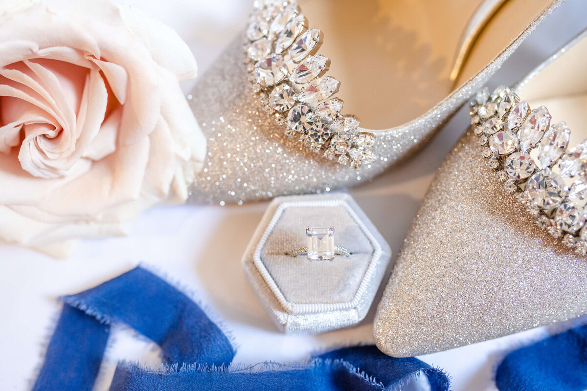 Flatlay of wedding details including shoes, flower, ribbon, ring box, and ring.