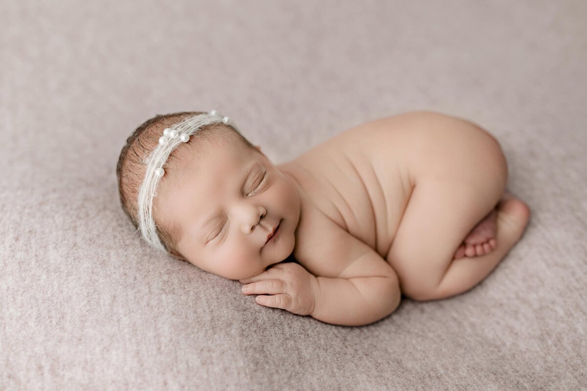 Newborn baby girl is wearing a headband and gently posed on a purple background layer with her little bum up