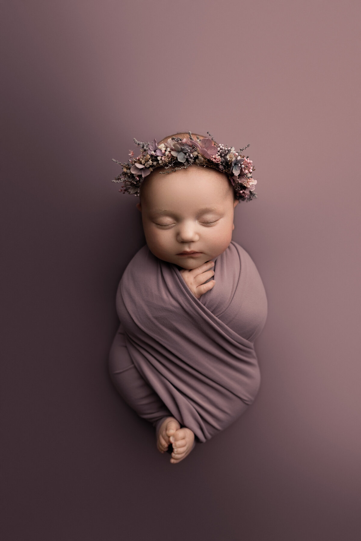 Main Line's best newborn photographer Katie Marshall captures an aerial image of a sleeping baby girl. Baby girl is swaddled in a dusty purple swaddle with her fingers and toes peeking out. She is wearing a coordinating floral crown and laying atop of a dusty purple stretch fabric.