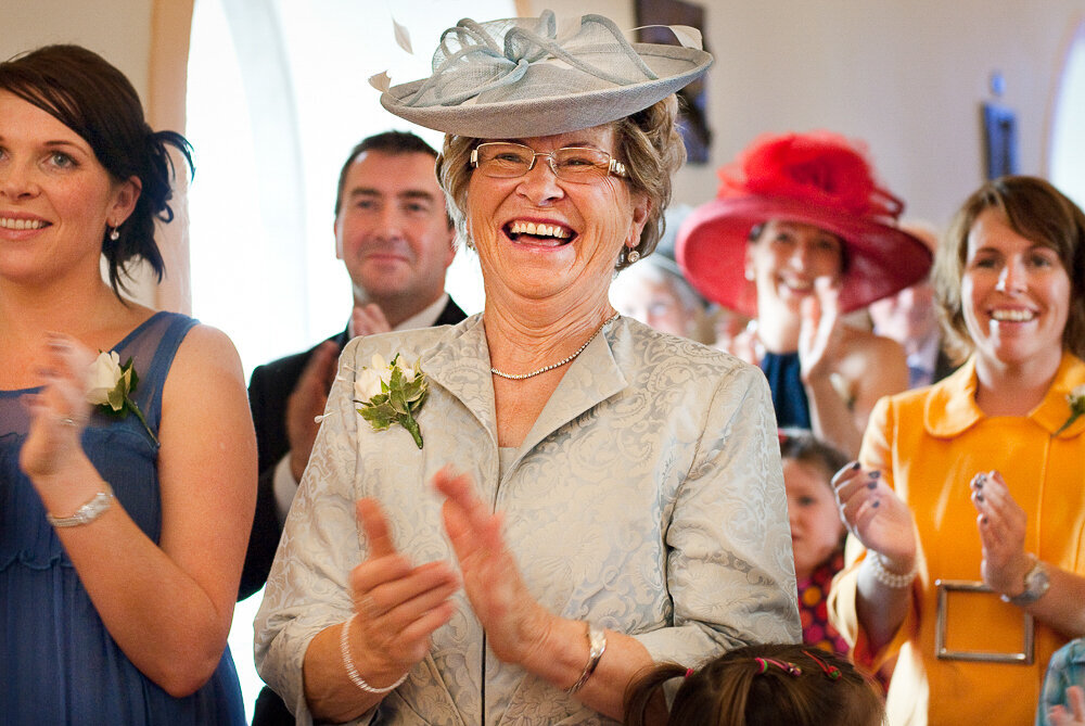 mother of the bride wearing a light, sage green outfit with matching hat clapping and laughing during wedding ceremony
