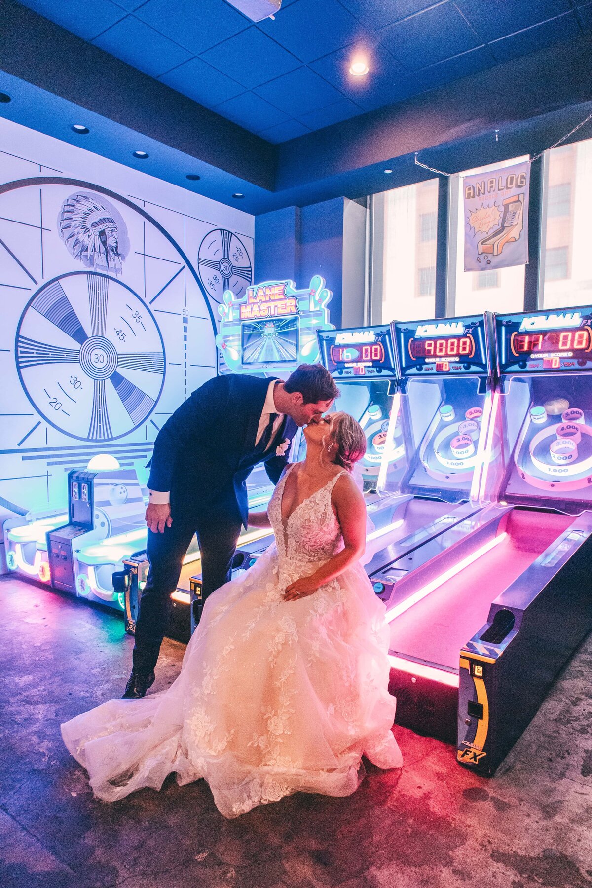 A newlywed couple kissing in an arcade, surrounded by colorful game machines and neon lights at a park farm winery wedding.
