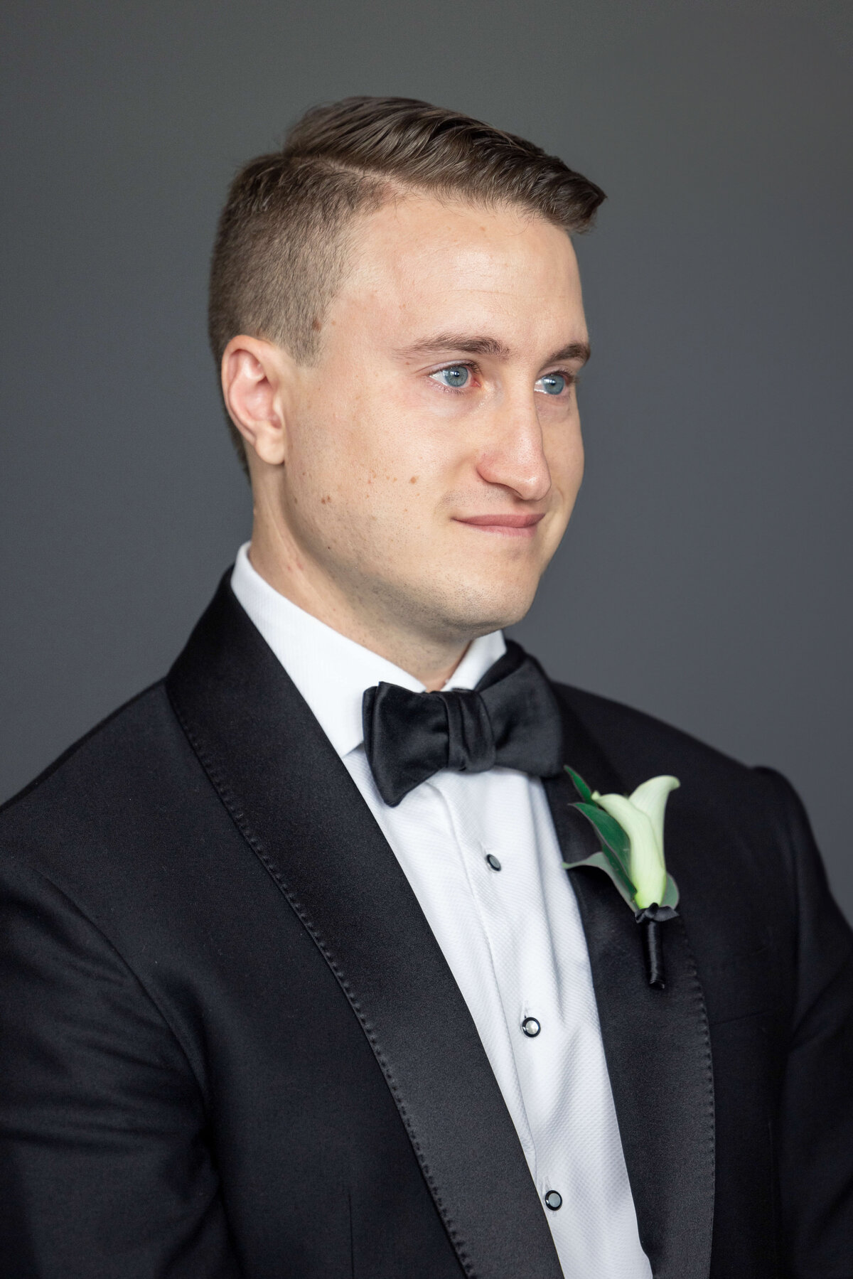 A man in a formal black tuxedo with a bow tie stands against a gray background. He has short hair and is wearing a white dress shirt. A white flower is pinned to his lapel.
