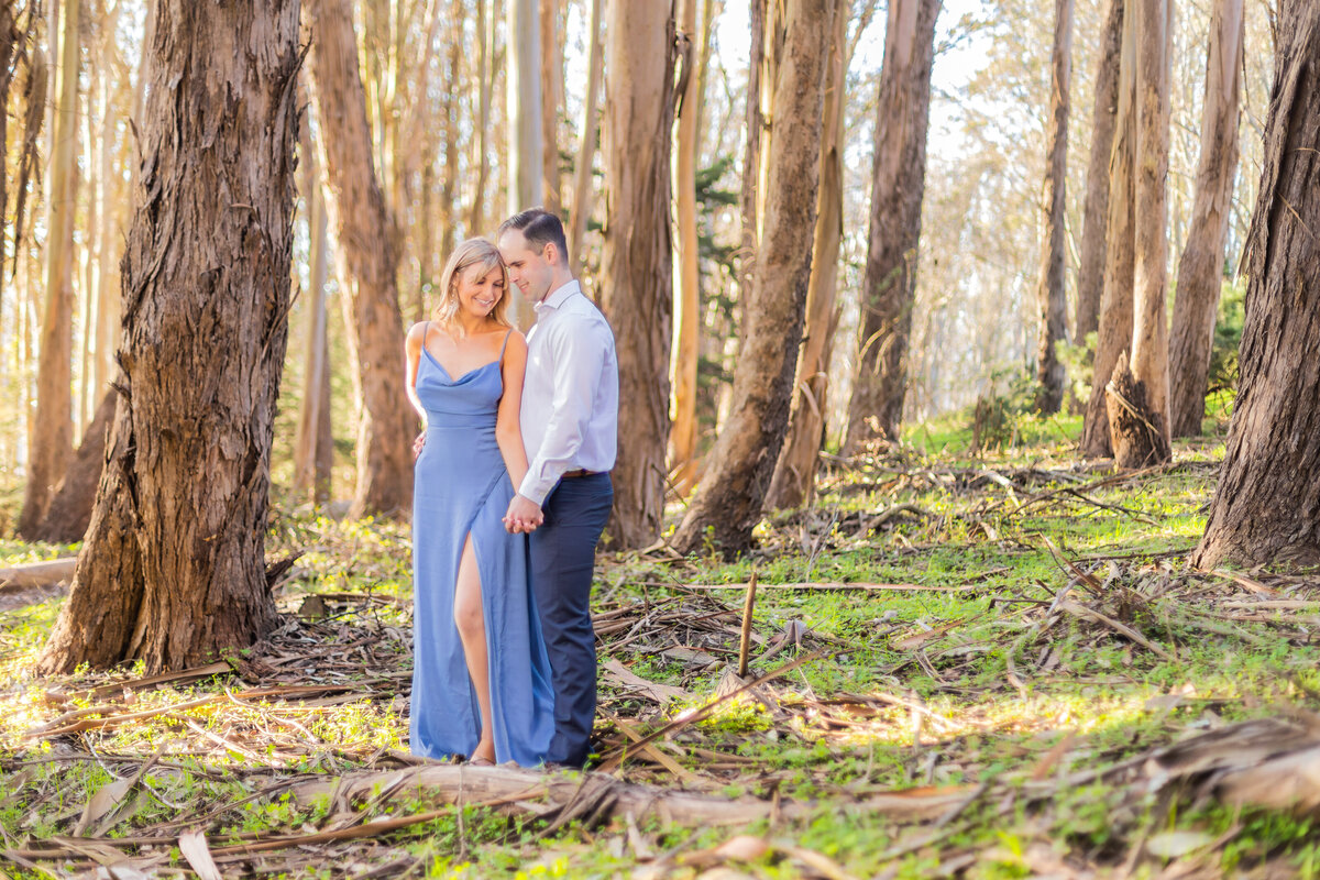 Engaged couple stand with the man behind the woman in the middle of the woods holding hands and smiling. Photo by wedding photograph, philippe studio pro.